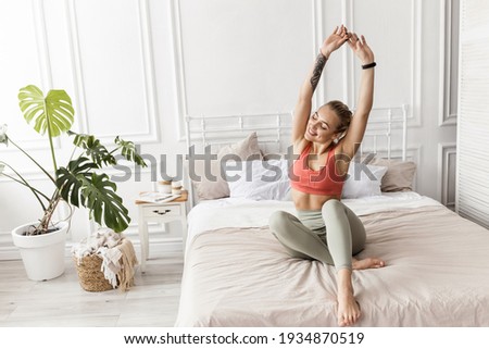 Happy blonde-hair woman is sitting on the bed, stretching after sleep, smiling. Attractive young female in stylish sportswear warm up before workout, morning training concept. High quality photo