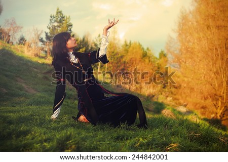 A beautiful young woman with dark hair and a historical dress posing on a meadow in open lanscape with a gesture of connection between heavenly and earth worlds
