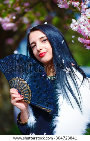 Beautiful Woman with flowers, glamour white fur and black fan in hand, posing next to blooming magical spring rosa sakura flowers. Flower background.