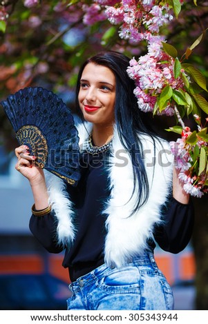 Beautiful Woman with flowers, glamour white fur and black fan in hand, posing next to blooming magical spring rosa sakura flowers. Flower background.
