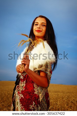 Young woman with ornamental dress and white fur standing on a wheat field with sunset. Natural background.