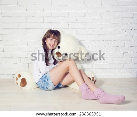 Charming girl sitting with her big white Teddy bear leaning against white brick wall. Studio photo