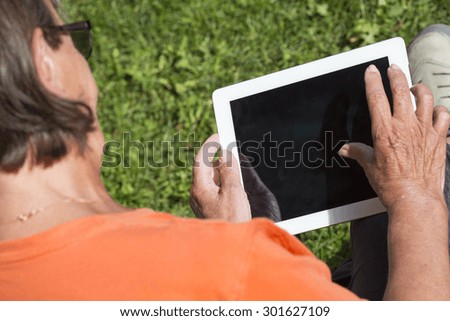 An elderly woman browsing the internet with a tablet outdoor with a green grass in the background.