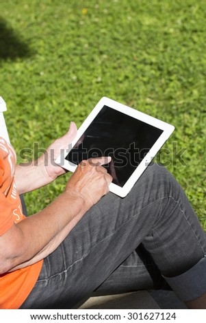 An elderly woman browsing the internet with a tablet outdoor with a green grass in the background.