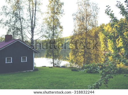 A red cabin or sauna by the lake.  Image taken during early autumn and sunset. An apple tree is in the right side and focus point is on the red cabin. Image has a vintage effect applied.