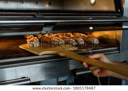 Close up of baker is taking off from oven the french baguette bread with wood peel at baking manufacture factory. French baguette bread bakery factory concept.