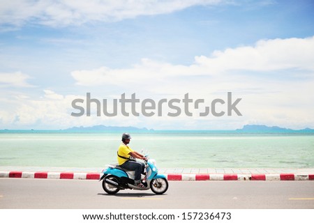 Man driving a motorcycle along the road, blue sea-sky background.