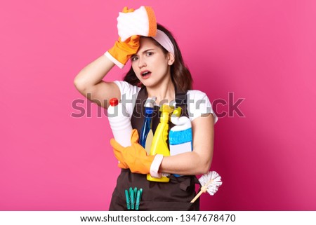 Young tired housewife in brown apron and white casual t shirt, with cleaning deretgents, isolated on pink background. Female has exuasted facial expressions, poses in photo sudio. Hygiene concept. Imagine de stoc © 