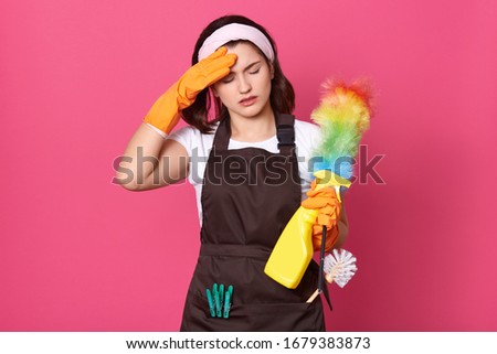 Close up portrait of tired housewife dresses brown apron, white casual t shirt and hair band, posing with cleaning deretgents isolated over pink background, having exuasted facial expressions. Imagine de stoc © 