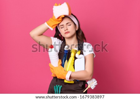 Indoor shot of young tired housewife wearing brown apron, white casual t shirt, haedband, posing with cleaning deretgents isolated over pink background, maid touching her forehead, holds sponge. Imagine de stoc © 