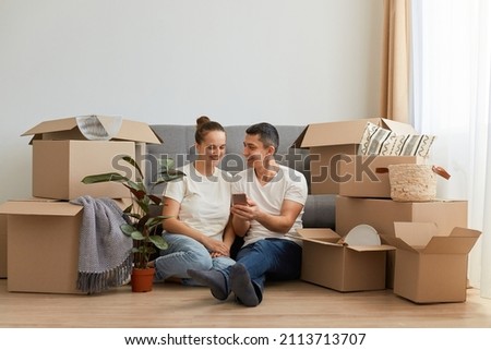 Indoor shot of young couple in love moving in a new apartment, sitting on the floor near, planning to redecorate their new home, man holding smart phone, showing ideas to his wife. Imagine de stoc © 