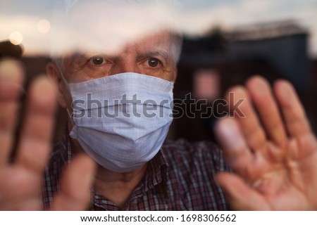 Elderly caucasian man wearing hand made protective face mask,in nursing care home,looking outside window, sadness,stress  hope in his eyes,self isolation due to global COVID-19 Coronavirus pandemic