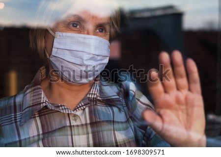 Elderly caucasian woman wearing hand made protective face mask, in nursing care home, looking outside window with sadness in her eyes, self isolation due to the global COVID-19 Coronavirus pandemic