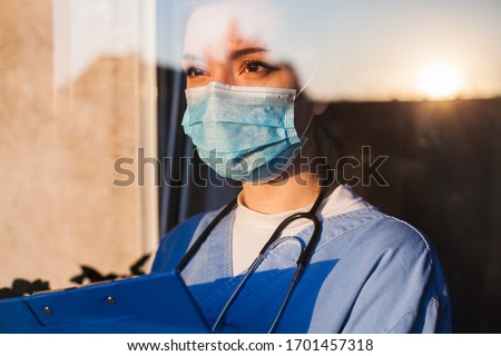 Young sad female caucasian UK US GP EMS doctor carer looking through ICU window,fear uncertainty in eyes,wearing face mask gazing at sun,hope faith in overcoming Coronavirus COVID-19 pandemic crisis  商業照片 © 