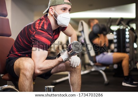 Young men working out wearing face mask  latex rubber gloves,performing bicep curl with dumbbells,COVID-19 pandemic social distancing rules while working out in reopened indoor gym,prevent  protect