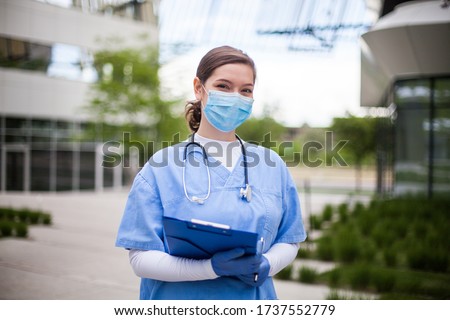 Female doctor holding blue clipboard standing outside hospital or clinic,frontline key medical worker portrait in modern care facility or nursing home complex yard,Coronavirus pandemic outbreak crisis