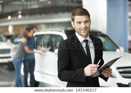 Let me assist you in your vehicle search. Handsome young classic car salesman standing at the dealership with the customers on the background