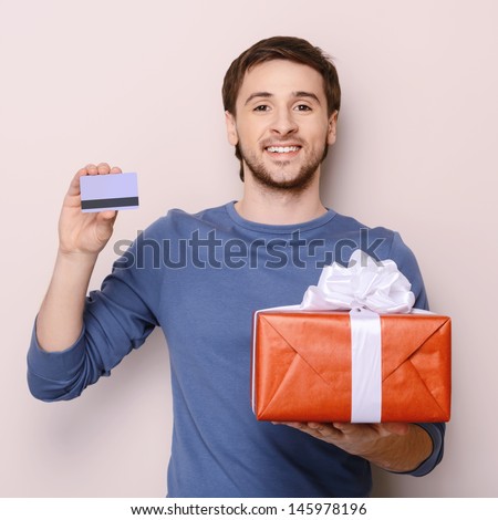 Portrait of young man holding gift box and a credit card. Handsome young smiley man holding a credit card in one hand and a gift box in another one