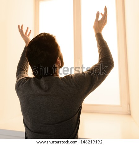 Waiting for miracle. Rear view of man standing in front of the window with his hands raised up