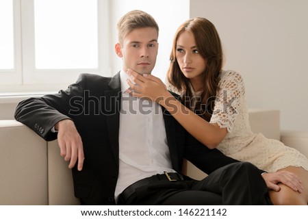 Serious couple. Beautiful young couple sitting on the couch while men in formal wear looking at camera and holding his hand on a girlfriend leg