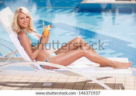 Beauty with cocktail. Attractive young women in bikini lying on the deck chair near the pool and holding a cocktail in her hand