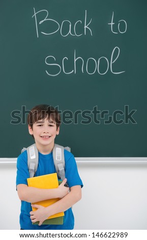 Welcome back to school! Cheerful little schoolboy holding the books and smiling while standing in front of blackboard