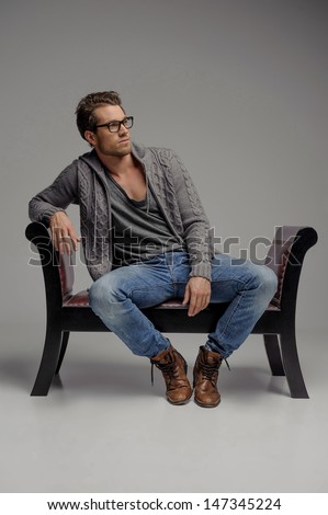 Thinking of the future. Handsome young men in glasses sitting on the chair and looking away while isolated on grey