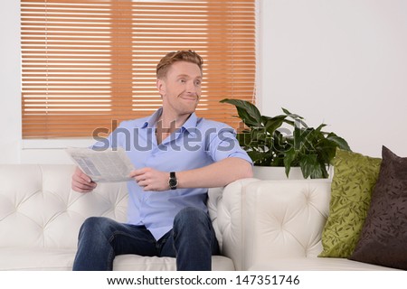 Reading a newspaper.  Cheerful young men reading paper and smiling while sitting on the couch