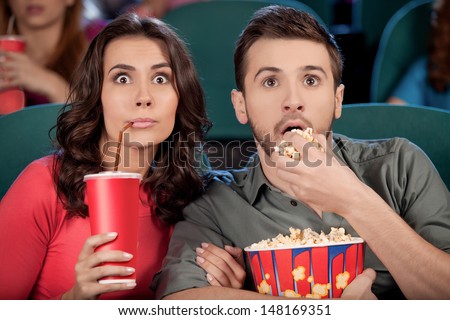 Exciting movie. Shocked young couple eating popcorn and drinking soda while watching movie at the cinema