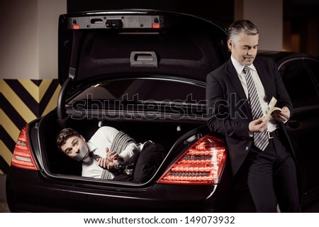 Kidnapped man. Tied up businessman lying in the car trunk and looking at camera while kidnapper counting money near him