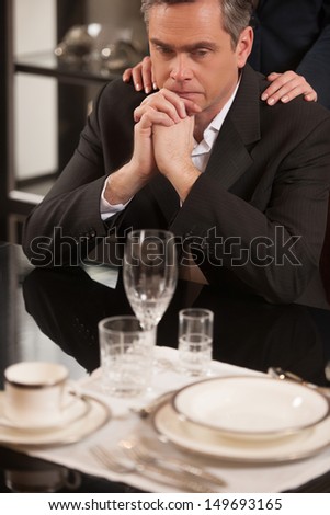 Depressed businessman in restaurant. Disappointed mature man in formalwear sitting at the restaurant and holding his hands on chin while woman holding her hands on his shoulders