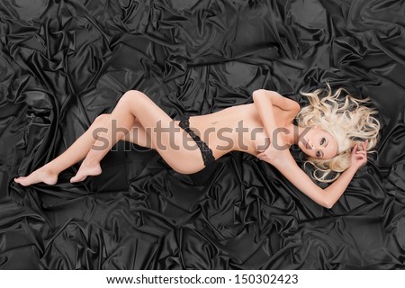 Naked and beautiful. Top view of beautiful topless woman covering her breast with hand and looking at camera while lying down on black cloth