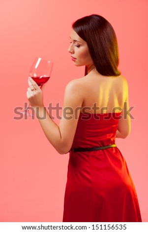 Woman with glass of wine. Rear view of beautiful young woman in red dress holding a glass with wine while standing isolated on colored background