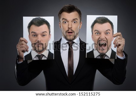Choosing mask on today. Surprised young man in formalwear holding two photographs of himself expessing different emotions while isolated on grey