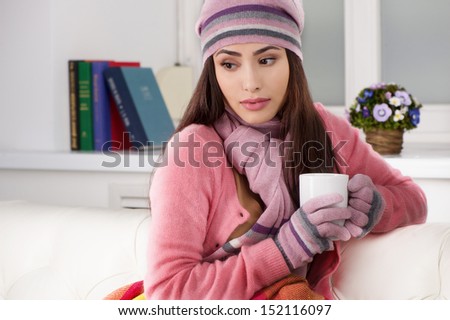 Girl in winter clothing. Beautiful young woman in hat and gloves drinking tea while sitting at home