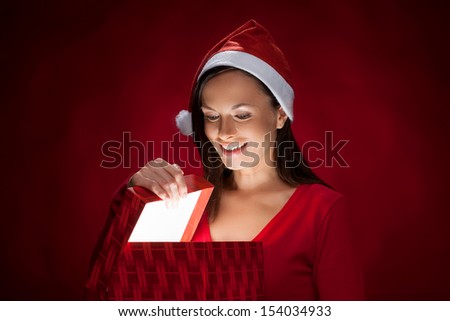 Christmas gift. Beautiful young woman in Santa hat holding a gift box and looking inside of it while isolated on red