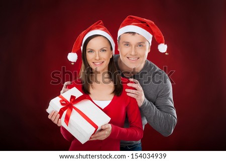 Together at Christmas Eve. Cheerful young couple in Santa hat holding a gift box and smiling while standing isolated on red