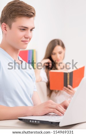 Loving couple at home. Cheerful young man working at the computer while his girlfriend reading book