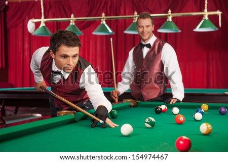 Pool game. Confident young man aiming the billiard ball with cue while his opponent smiling on the background
