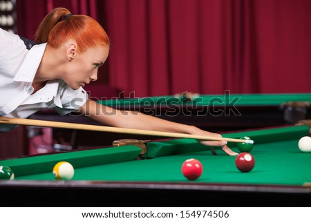Woman playing pool. Side view of confident young woman aiming the billiard ball with cue
