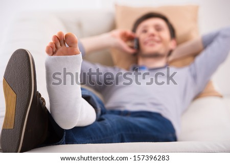Man is laying on sofa and talking by phone with bandage on leg