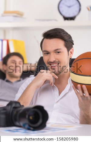 Man holding basketball ball and looking at camera. Another man is resting on background