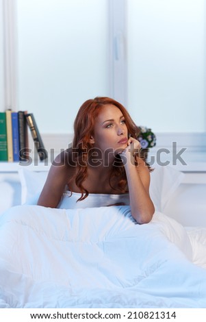 Beautiful woman with depression sitting in bed. Sad red-haired woman sitting on her bed and thinking