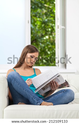 Attractive girl reading magazine on sofa. girl holding magazine at home next to open window