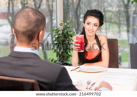 Smiling attractive woman drinking juice with straw. back view of man listening to girl at lunch break