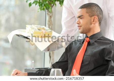man sitting in restaurant and looking at money. waiter holding tray full of money