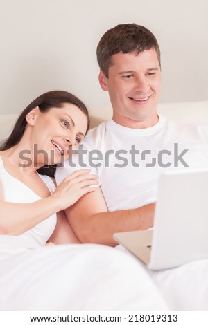 Couple using computer together in bed. Beatiful lovers relaxing lying on their bed