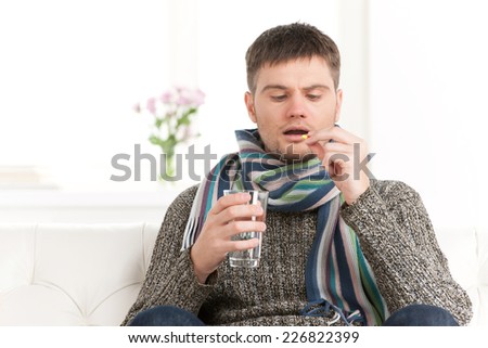 Sick caucasian man holding pills and water. Sick young man with flu drinks water on sofa