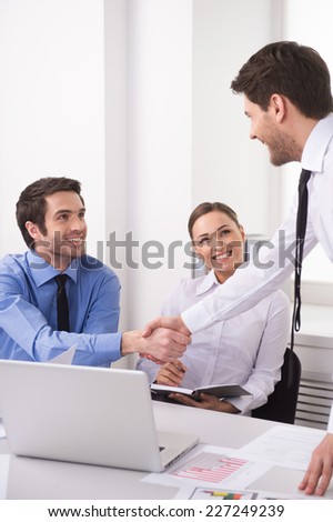 Side view of three people working on computer at office. Businesspeople sitting at desk in office and shaking hands