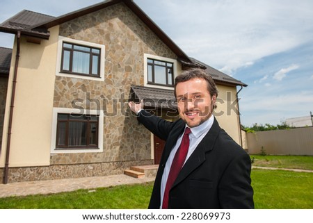 closeup on smiling real estate agent ready to sell house. Male real etate agent in front of home pointing to house